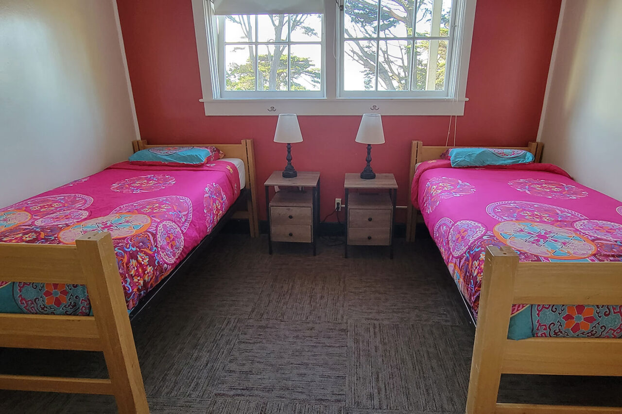 two freshly made twin-sized beds with pink bedspreads in a two-person private room at HI San Francisco Fisherman's Wharf hostel. The wall behind the beds is a light red color, and there is a large window overlooking the trees.