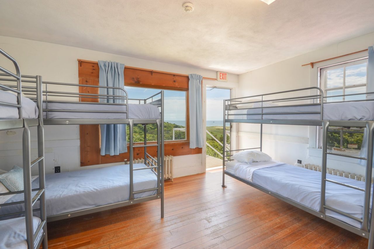 a large and airy dorm room at HI Truro hostel on Cape Cod. There are three sets of freshly made bunk beds with hardwood floors and large windows looking out over the landscape.