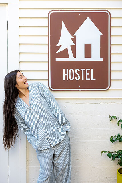 a young woman with long black hair wearing green-striped pajamas stands in front of a white building while looking at a brown hostel sign.