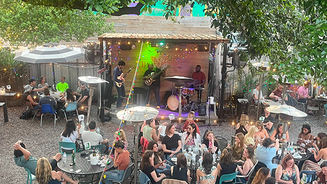 A view from overhead of Bachannal in New Orleans. A crowd of people sit at picnic tables under the trees and a live band plays on a small stage in the background.