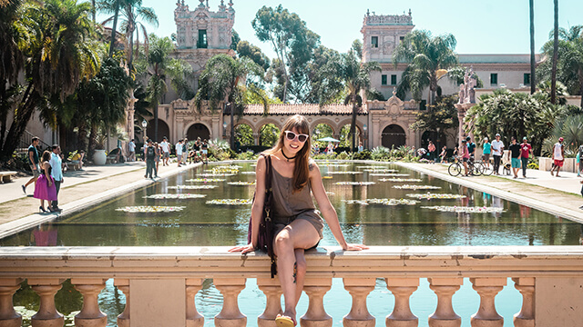 a woman sitting in front of a large Mission-style building with a pond at Balboa Park in San Diego