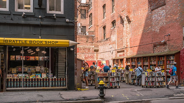 Brattle Book shop in Boston. The shop's sign is a giant yellow pencil, and there are people browsing outdoor shelves of books in the alleyway