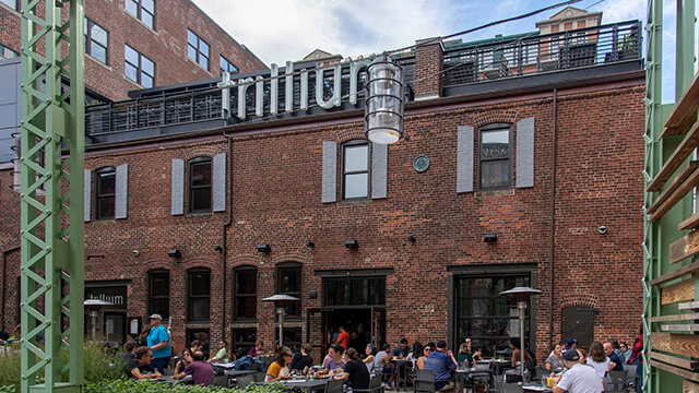 a large brick building with a sign reading "trillium" at the top. There are lots of people sitting at tables outside in front of the building. 