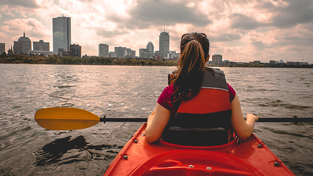 a rear view of a kayaker with a red life vest and ponytail. She is holding an oar and overlooking the Boston skyline.