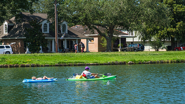 a kayaker paddles down Bayou St. John in New Orleans, pulling a child on a raft behind him. On the shore, two people practice boxing in front of a line of houses.