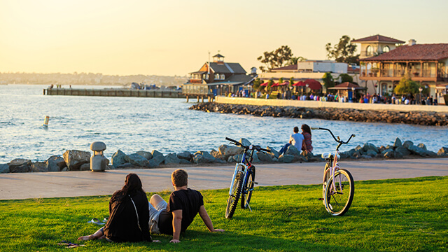 Two travelers sit next to bicycles on a small green lawn overlooking the water and San Diego's Seaport Village