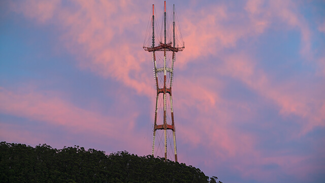 a sunset with pink clouds and a purple sky behind a large red and white tower on top of a large hill covered in eucalyptus trees in San Francisco