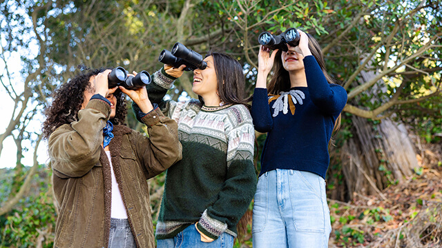 Three young travelers in sweaters stand outside in the trees, each holding a pair of binoculars to her face and looking in a different direction.