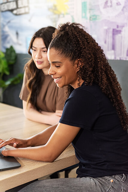 two young women sit at a long table in a hostel looking at a laptop together