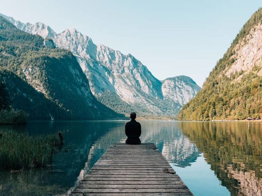 silhouette of a person sitting alone at the end of a dock on a mountain lake