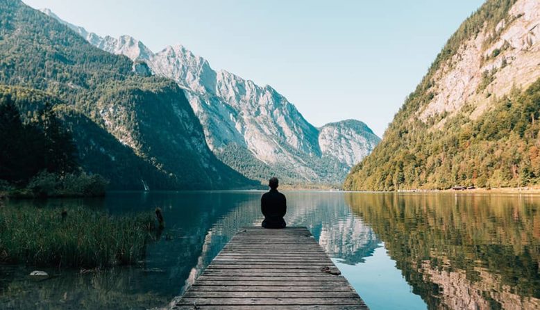 silhouette of a person sitting alone at the end of a dock on a mountain lake