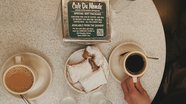 an overhead view of a small round table at cafe du monde in new orleans. In the center of the table is a plate full of fresh beignets. On either side is a cup of coffee.