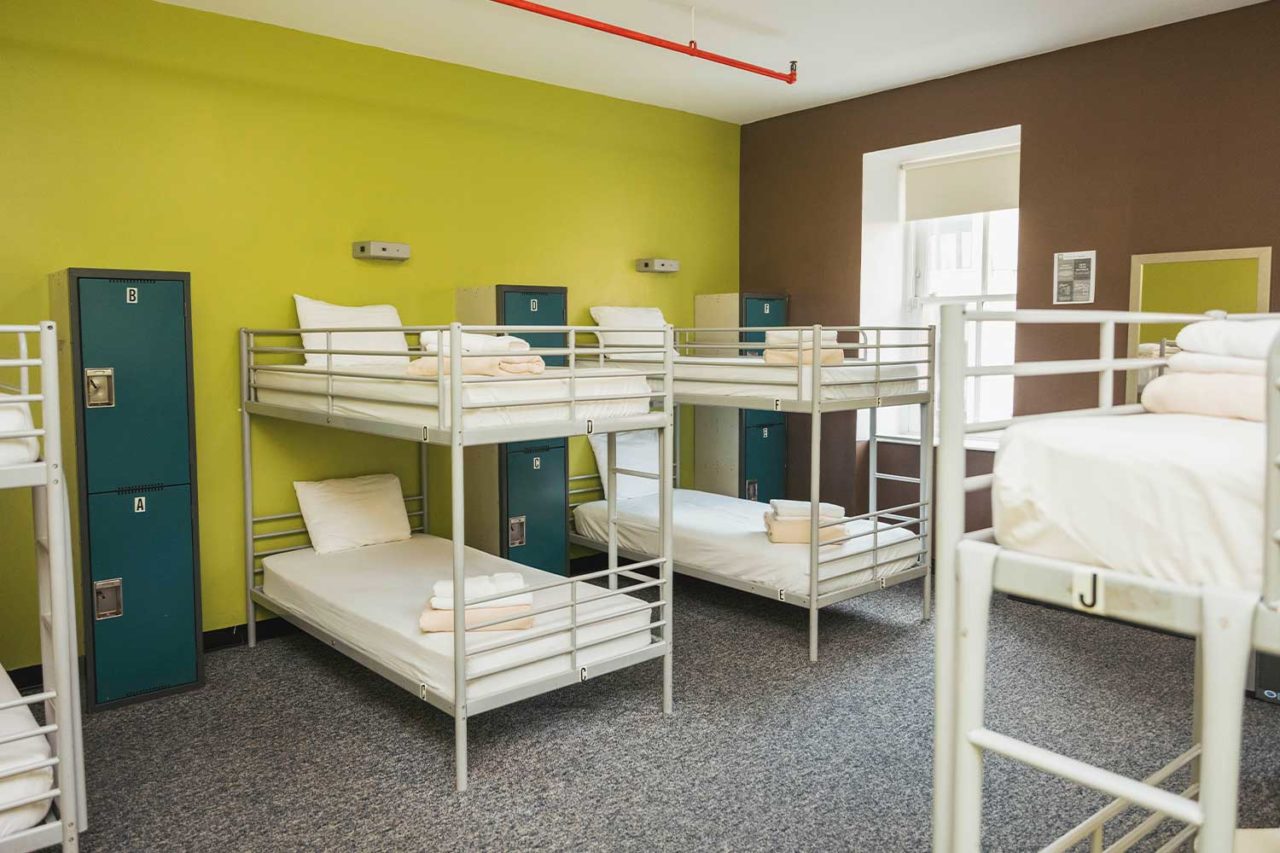 a large dorm room at HI New York City hostel with bright green walls, grey carpet, and four sets of bunk beds. The beds are made up with crisp white linens. Along the back wall, there are individual secure lockers for guests' belongings.
