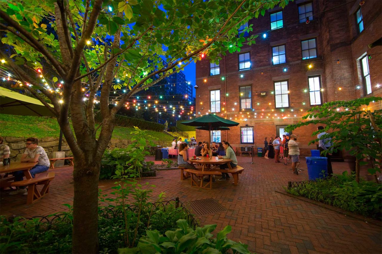 The large outdoor brick patio at HI New York City hostel at night. The patio is surrounded by trees strung with yellow cafe lights. There are picnic tables with umbrellas and benches, with people sitting outside, on the patio.