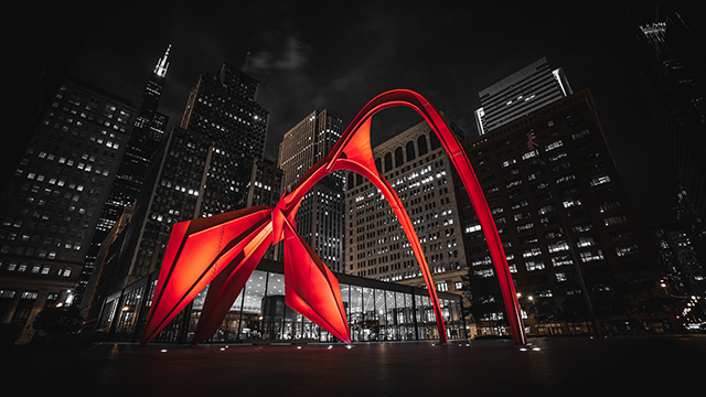 a giant red sculpture in front of tall illuminated buildings seen at night