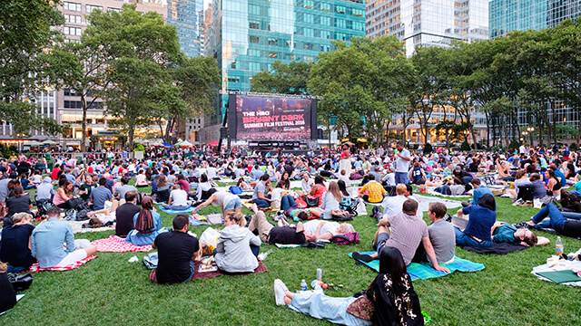 a crowd of people sitting on a green lawn in front of a large outdoor movie screen with skyscrapers in the background