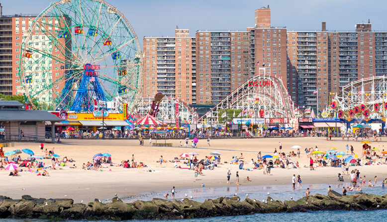 A view of the beach at Coney Island in Brooklyn, with a large ferris wheel, roller coaster, and housing along the beach