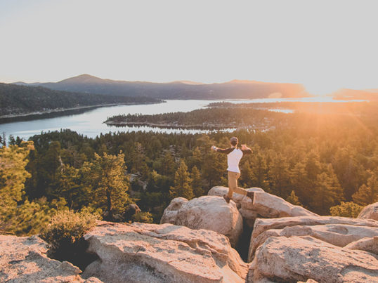 a hiker jumps between two boulders with trees and a lake in the background