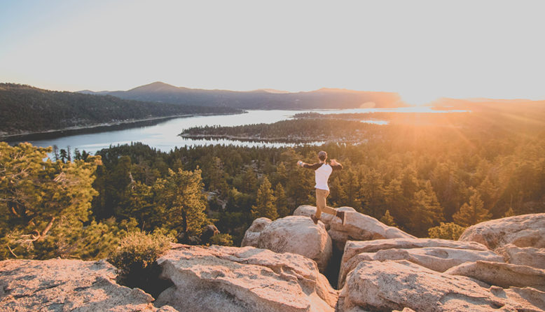 a hiker jumps between two boulders with trees and a lake in the background