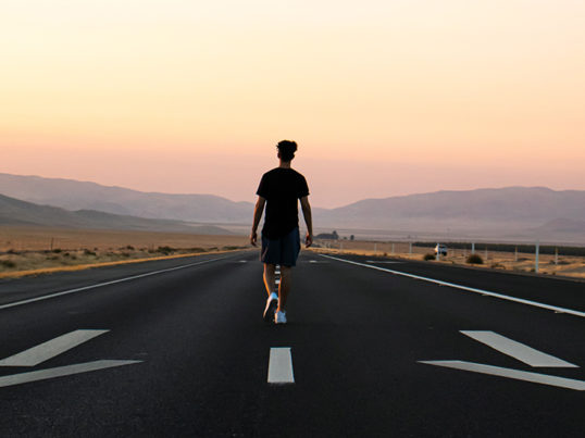 a person with his back to the viewer walks in the middle of a road with arrows pointing the other direction and the sunset in the background