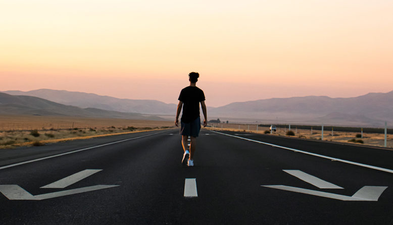 a person with his back to the viewer walks in the middle of a road with arrows pointing the other direction and the sunset in the background