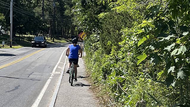 a man in a blue shirt rides his bicycle past green foliage on a bicycle trail in hyannis massachusetts