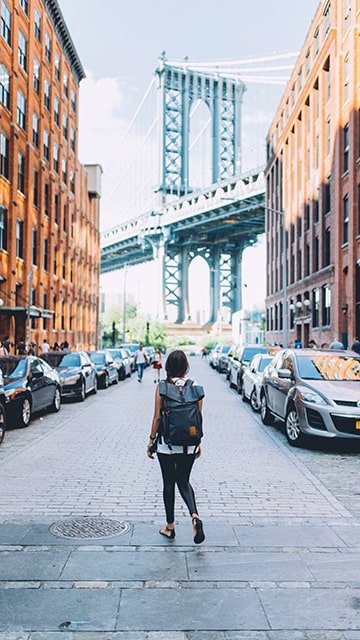 a woman with a black backpack stands alone in the street between red brick buildings looking at a blue bridge