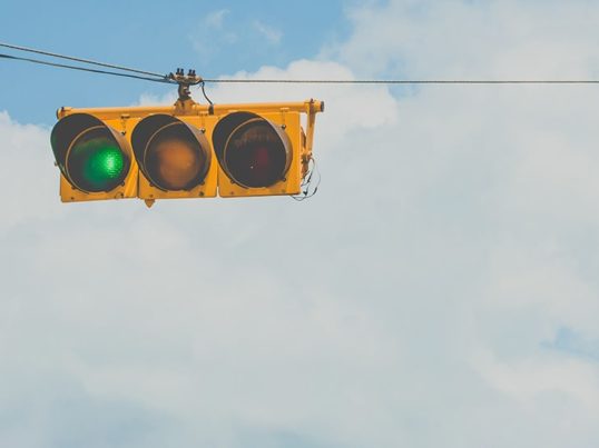 a horizontal traffic light with the green illuminated against a background of clouds