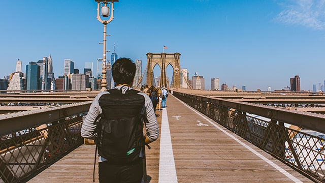 a man in a grey sweatshirt with a black backpack stands on the brooklyn bridge