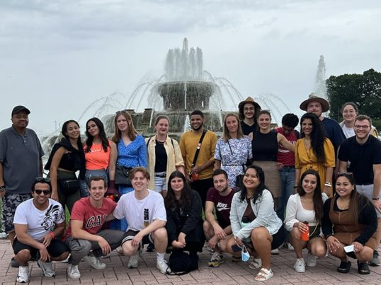 a large group of young people from all over the world stand in front of a large outdoor fountain