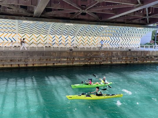 two colorful kayaks paddle through turquoise water below a bridge