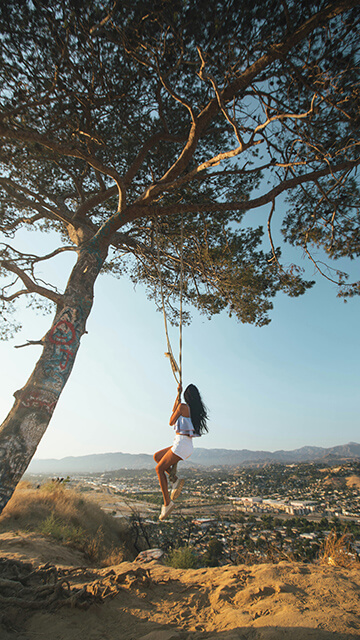 a young woman with long dark hair wearing white shorts and a white top swings on a rope swing in Elysian Park overlooking Los Angeles