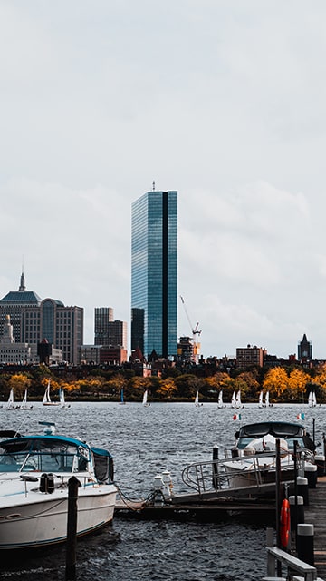 a tall glass skyscraper sits on the banks of the Charles River with boats and autumn trees in the foreground