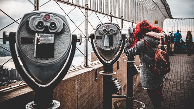 a woman with red hair looks out of a viewfinder from the top of a building