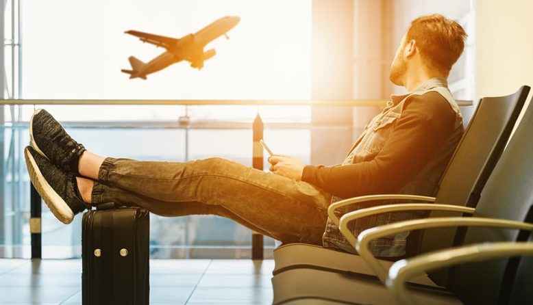 a man sits in a waiting area at an empty airport with his feet up on a suitcase while watching a plane take off