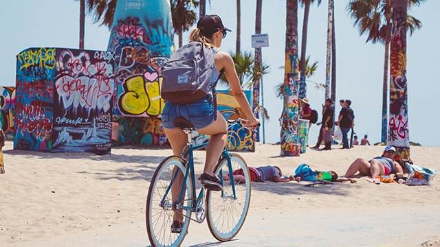 a woman wearing shorts and a backpack rides a bicycle on the beach in front of graffiti and palm trees