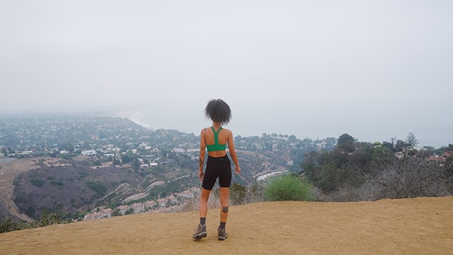 a woman in hiking boots stands on a hill overlooking the los angeles skyline