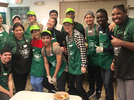 a group of young volunteers in green aprons smile while posing in a food pantry kitchen