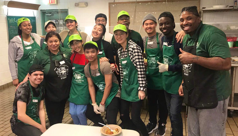 a group of young volunteers in green aprons smile while posing in a food pantry kitchen