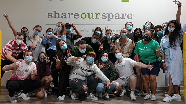 a large group of young volunteers pose in front of a wall painted with the Share Our Spare organization logo