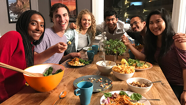 a diverse group of six young people sit around a table covered with plates and bowls of colorful food