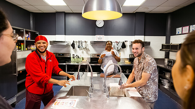 people stand around a large metal kitchen island laughing 