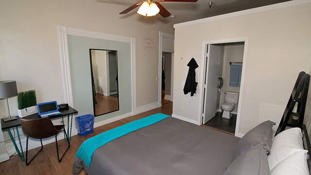 a full-sized bed with grey comforter in a room containing a desk with laptop and chair. A private bathroom with toilet and shower is visible through an open door.