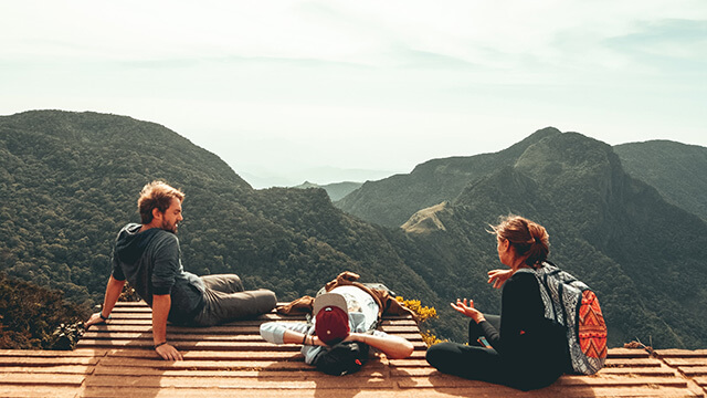 Three people in hiking clothes sit on a platform overlooking the mountains while having an animated discussion