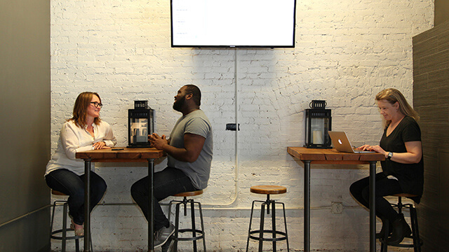 two people talk at a high-top table while a woman works on a laptop at the table next to them