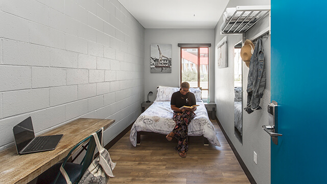 a woman sits at the foot of a bed reading in a small room with a desk and laptop