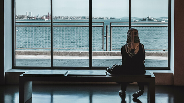 a woman sits on a bench with her back to the camera while looking out a large window onto the water