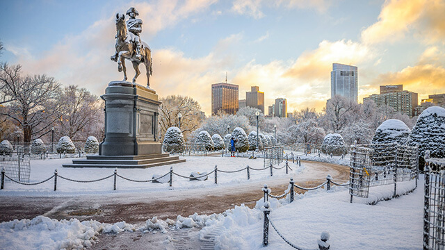 a statue of a person on a horse on snow-covered ground in the daytime with the boston skyline buildings in the background