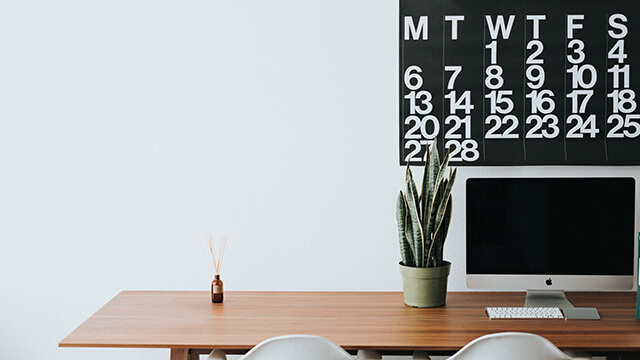 a desk with a computer and plant and a black and white calendar on the wall behind it