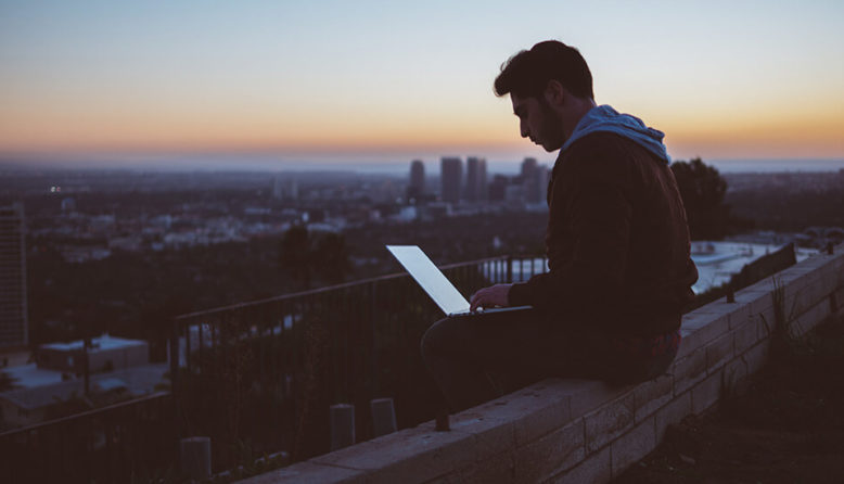 a man sits on a rooftop at sunset working on a laptop